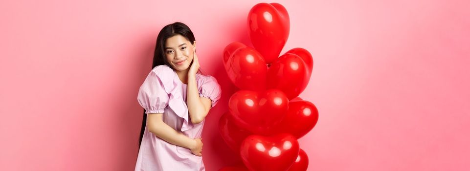 Beautiful asian girl in dress smiling coquettish, flirting on valentines day, looking sensual at camera, posing near valentines heart balloons, pink background.