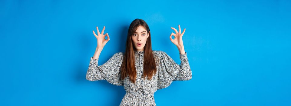 Excited beautiful woman in dress say wow, recommend cool thing, praising and looking impressed, standing on blue background.