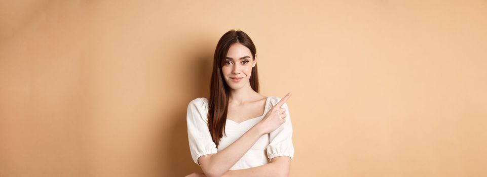 Smiling good-looking girl in white dress pointing aside, showing left logo and staring at camera confident, standing on beige background.