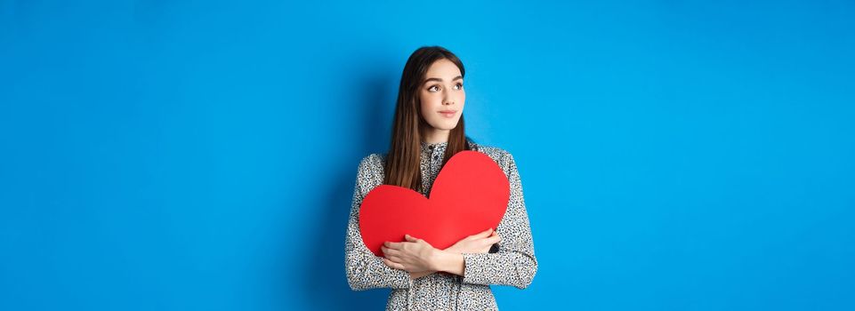 Valentines day. Romantic girl looking dreamy at upper left corner and smiling, holding big red heart cutout, standing on blue background.