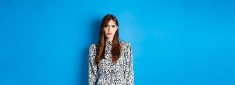 Moody frowning girl in dress looking at unfair thing, standing offended or upset against blue background.