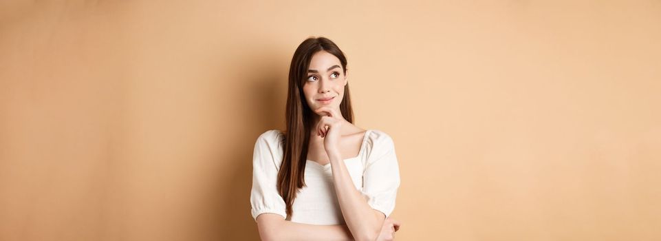 Very interesting. Pensive cute girl thinking and looking at upper left corner logo, smiling pleased, having good idea, standing on beige background.