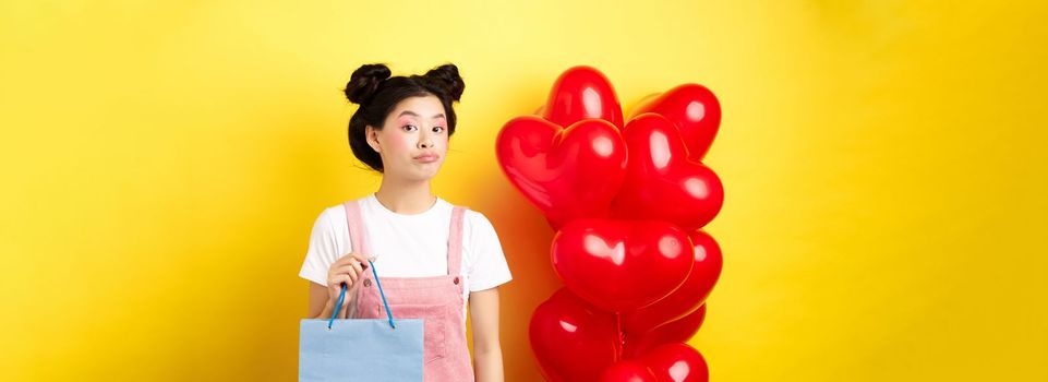 Happy Valentines day. Stylish asian single girl buying herself gift, holding shopping bag and looking unbothered at camera, standing over yellow background.