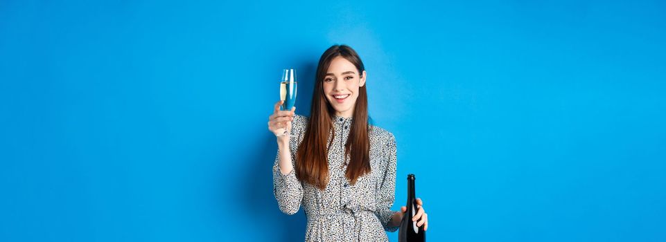 Celebration. Happy young woman in dress raising glass of champagne, holding bottle and smiling, having fun at party, standing on blue background.