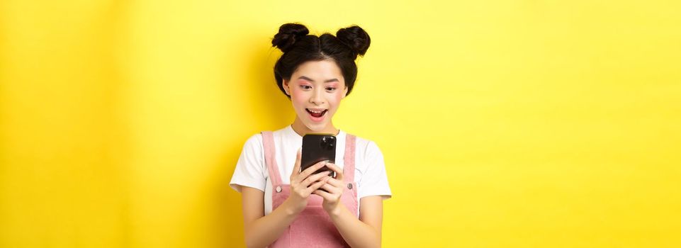 Pretty asian girl looking excited at screen, reading message on phone and smiling happy, standing in summer clothes on yellow background.