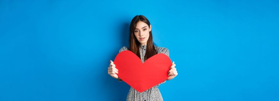 Valentines day and relationship concept. Tender young woman in dress stretch out hand and giving big red heart to you, making confession, standing romantic on blue background.