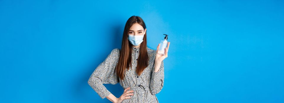 Covid-19, social distancing and healthcare concept. Beautiful girl in medical mask using hand saniziter, holding antiseptic bottle, standing on blue background.