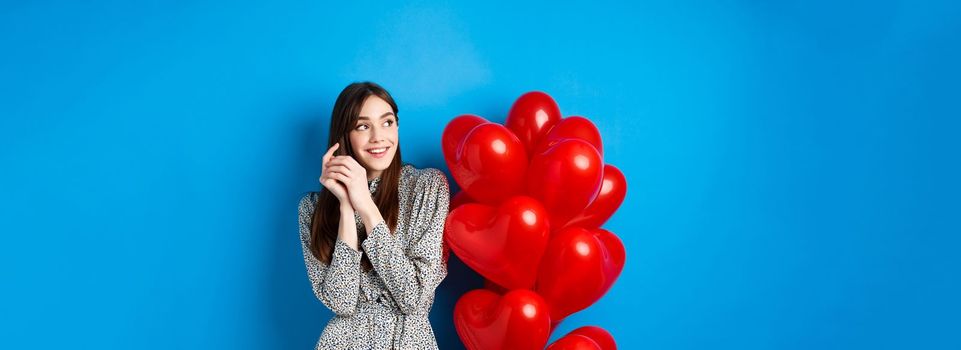 Valentines day. Romantic young woman in dress, looking dreamy left and smiling, standing near red hearts balloons, blue background.