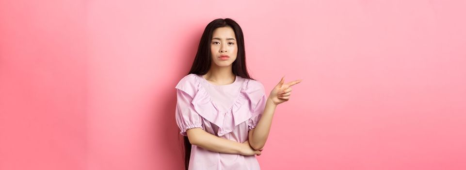 Skeptical chinese girl pointing finger left, looking unamused and serious, showing offer, standing in dress on pink background.