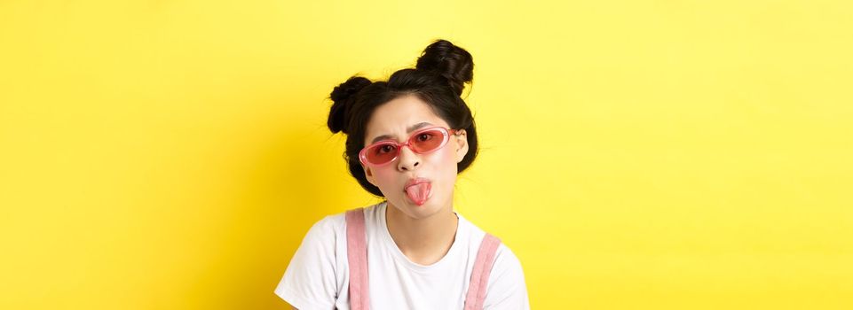 Summer and beauty concept. Childish asian girl in sunglasses showing tongue, being rude, standing on yellow background.