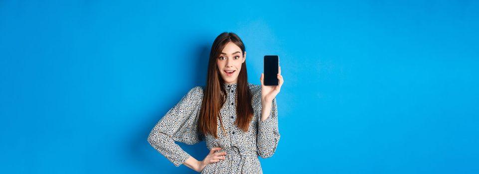 Amazed smiling woman showing empty smartphone screen, look impressed, recommending online promo, blue background.