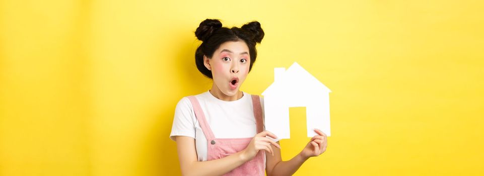 Real estate and family concept. Excited asian teen girl showing paper house cutout and gasping amazed, standing on yellow background.