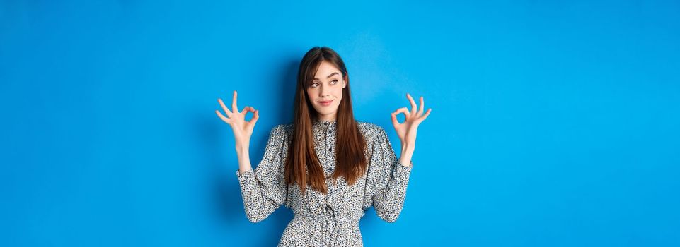 Not bad. Pleased smiling woman with long hair, wearing dress, showing okay signs and looking satisfied, praising good job, looking left at logo, blue background.