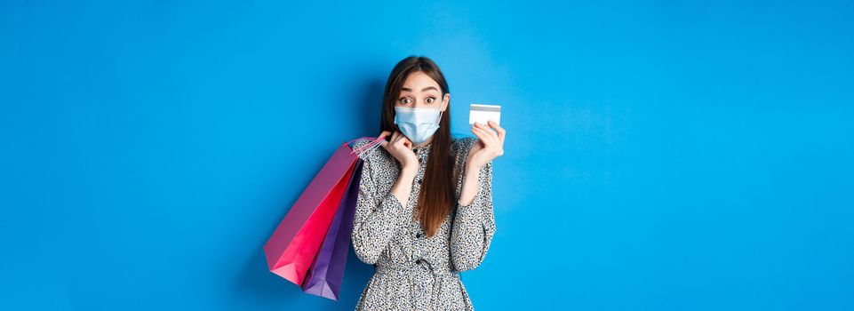 Covid-19, pandemic and lifestyle concept. Excited woman wearing medical mask while shopping, showing plastic credit card and holding bags, blue background.