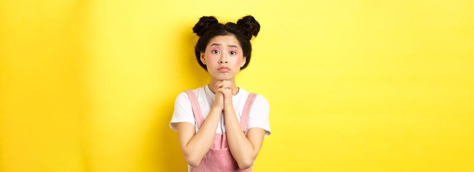 Hopeful asian stylish girl holding hands in begging sign, waiting with hope, asking for favour or help, standing on yellow background.