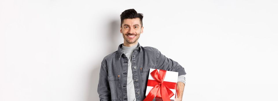 Happy Valentines day. Smiling handsome man bring gift on romantic date, holding box with present and looking romantic at camera, white background.