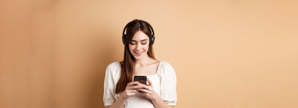 Smiling woman texting message on phone and listening music in wireless headphones, watching video on smartphone, beige background.