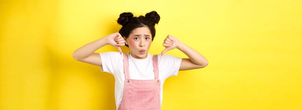 Nervous asian girl feeling bad, showing thumbs down and frowning sad, show negative emotion, standing in summer clothes on yellow background.