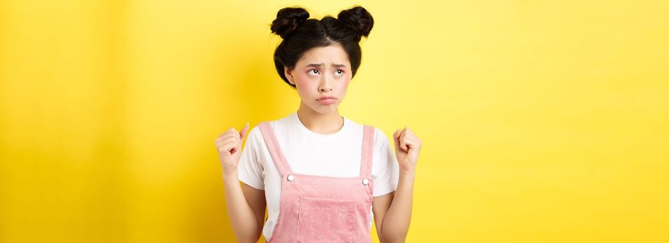 Timid angry girl sulking and clenching teeth, feeling mad but looking cute at upper left corner, standing with makeup on yellow background.