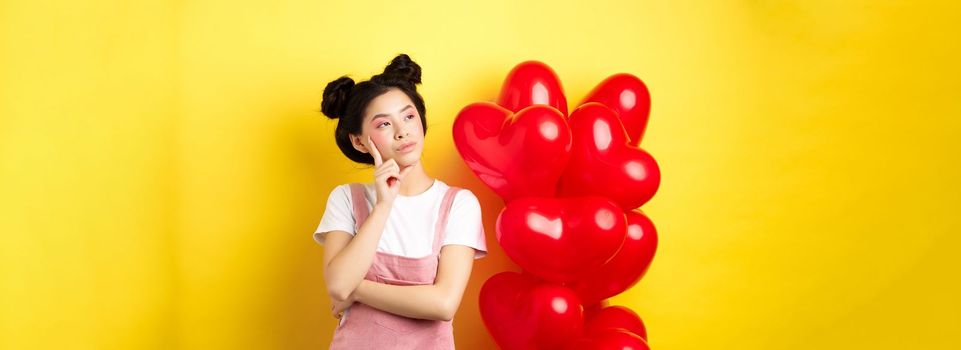 Valentines day concept. Pensive asian woman looking left, thinking about romantic date, standing near red hearts balloons on yellow background.