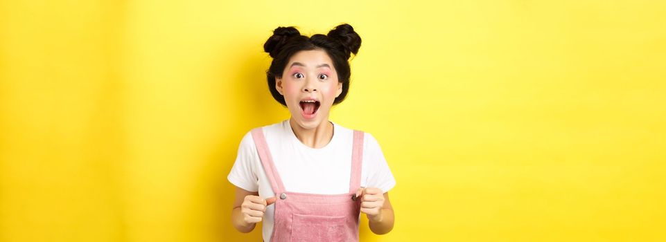 Excited asian woman open mouth, looking fascinated and amazed, receive good news, standing in summer clothes on yellow background.