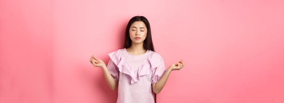 Stay calm. Peaceful asian woman meditating, hold hans in zen gseture and close eyes, practice yoga, standing patient on pink background.