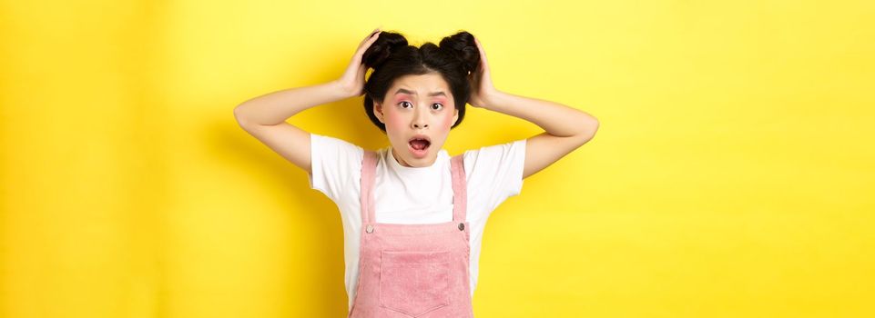 Shocked asian girl gasping worried, checking her hairstyle, standing anxious on yellow background.