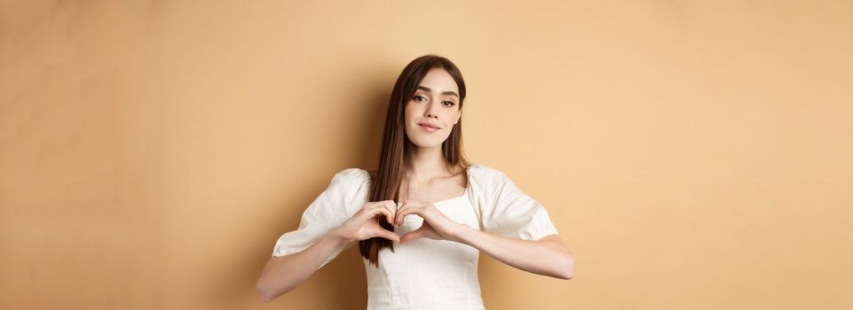 Valentines day and romance concept. Romantic caucasian girl in white dress showing heart gesture and smiling, say I love you, standing over beige background.