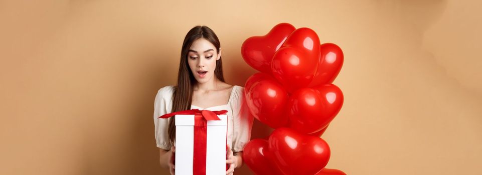 Surprised beautiful woman in romantic outfit, standing near heart balloons and looking at her gift on Valentines day, standing on beige background.