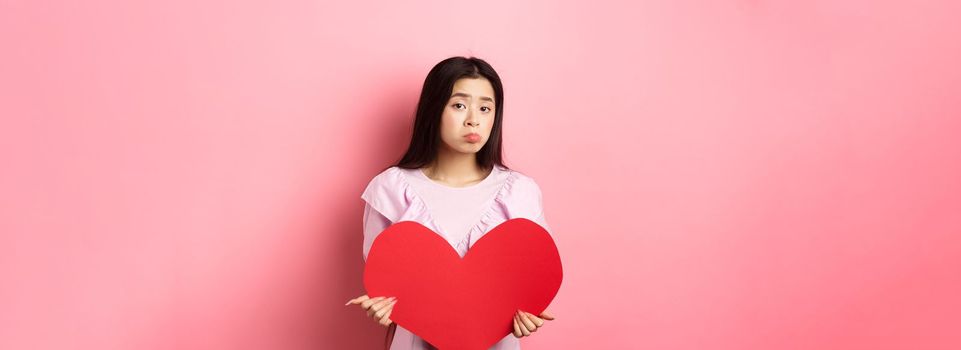 Valentines concept. Single teenage asian girl wants to fall in love, looking sad and lonely at camera, sulking distressed on lovers day, holding big red heart cutout, pink background.