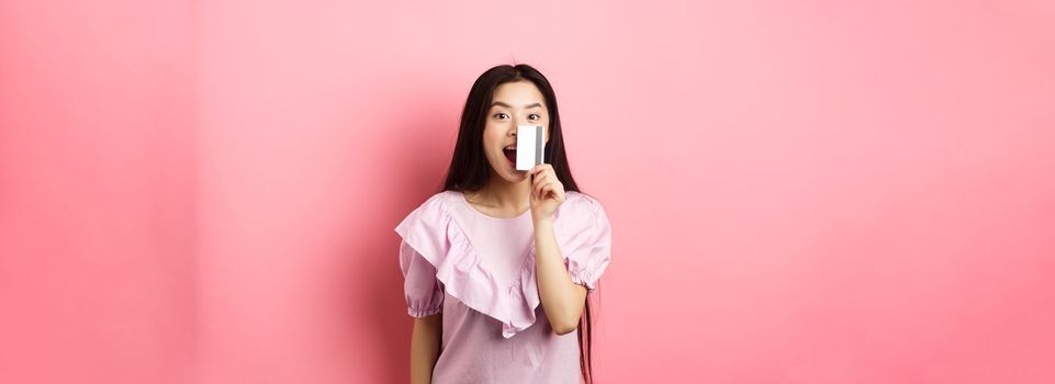 Excited asian girl showing plastic credit card and smiling, going on shopping, standing against pink background.