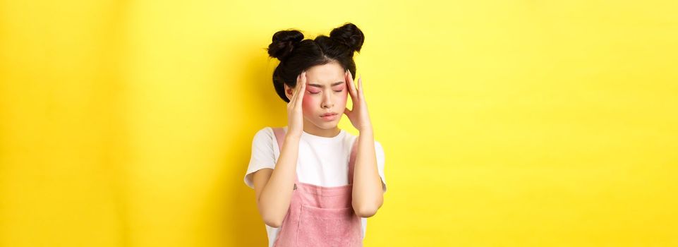 Image of stylish asian woman with bright makeup, having headache, touching head and looking tired, have painful migraine, yellow background.