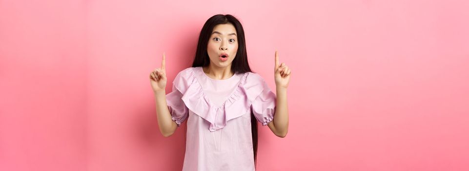 Excited asian girl showing awesome promo offer, pointing fingers up and gasping amazed, standing on pink background.
