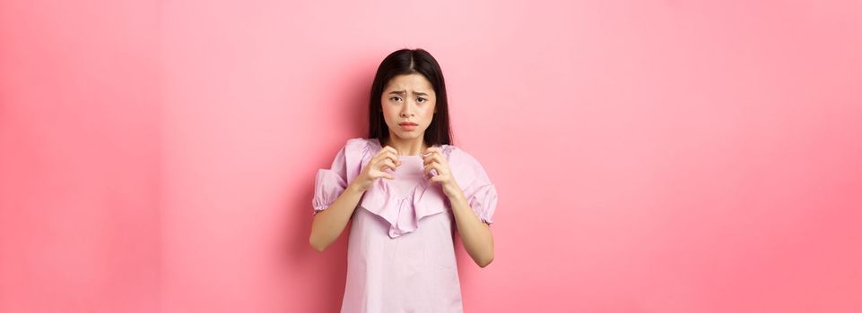 Scared asian woman victim, looking frightened, trembling from fear, stare at scary thing, standing anxious on pink background.