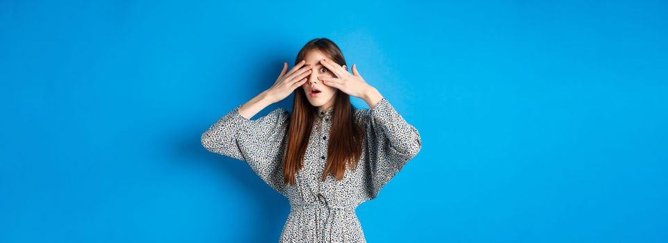 Intrigued young girl peeking through fingers with excitement, seeing interesting thing, standing on blue background.