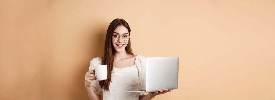 Cheerful woman in glasses drinking coffee and working on laptop, smiling at camera, standing on beige background.