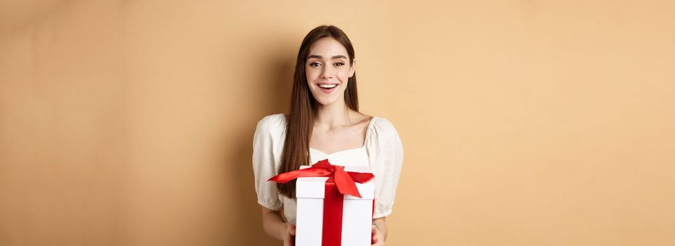 Happy valentines day. Cheerful smiling girl giving surprise gift to lover, looking at camera and holding present in box, standing on beige background.
