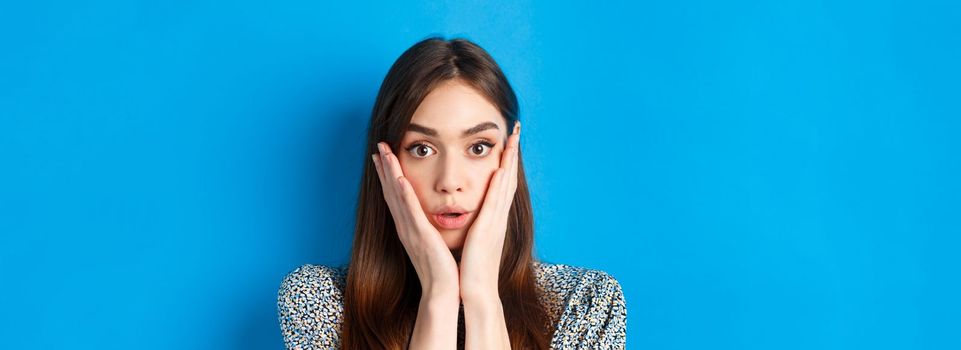 Close-up portrait of surprised cute woman holding hands on cheeks and gasping, stare at camera shocked, standing against blue background.