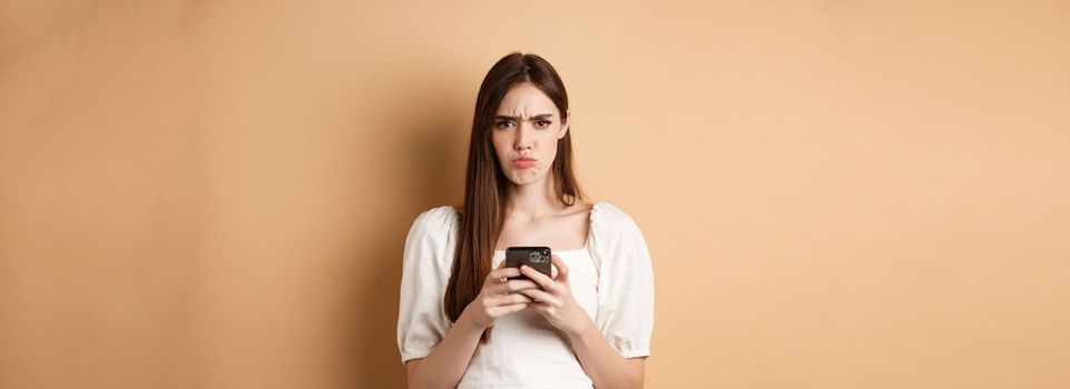 Disappointed girl with smartphone frowning, pucker lips upset, reading bad news on phone, standing on beige background.