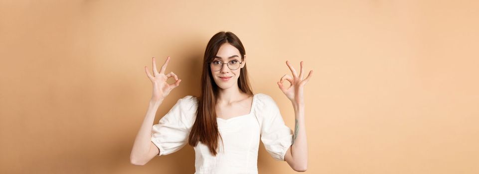 Satisfied female model in glasses show okay sign, looking pleased, agree and approve, standing on beige background.