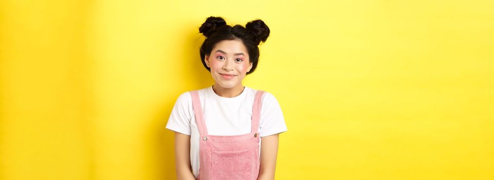 Cute asian woman with makeup and summer clothes, smiling silly and happy at camera, yellow background.