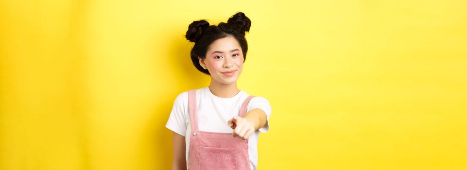 Stylish japanese girl with beauty makeup pointing at camera, smiling at you, beckon and invite people, standing on yellow background.