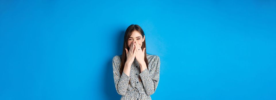 Cheerful caucasina girl in dress laughing and having fun, covering mouth with hands and chuckle over something funny, standing on blue background.