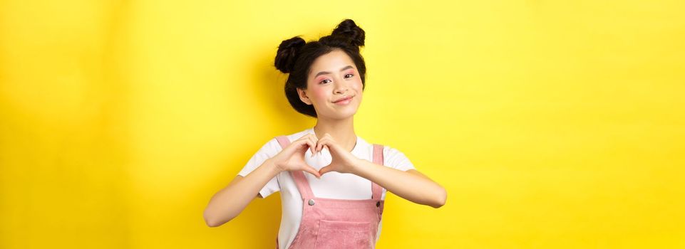 Valentines day. Cute asian girl sending her love, showing heart gesture and smiling at camera romantic, standing on yellow background.