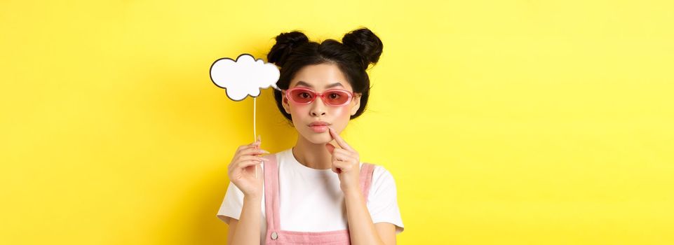 Summer and fashion concept. Stylish asian girl in sunglasses holding comment cloud on stick and looking thoughtful, thinking or saying something, standing on yellow background.