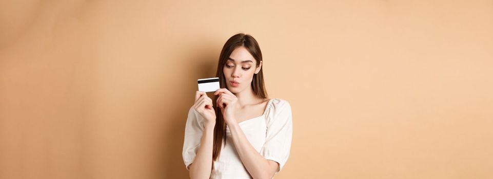 Cute young woman looking thoughtful at plastic credit card, thinking of shopping, standing on beige background.