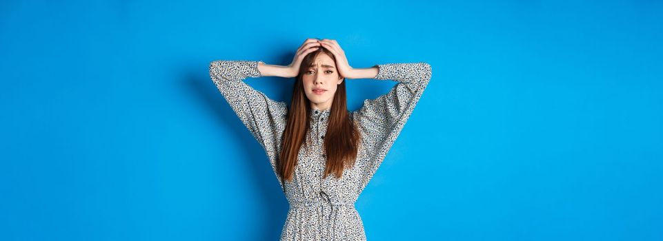 Frustrated adult woman with natural hair holding hands on head and panicking, looking alarmed and scared, standing in dress against blue background.