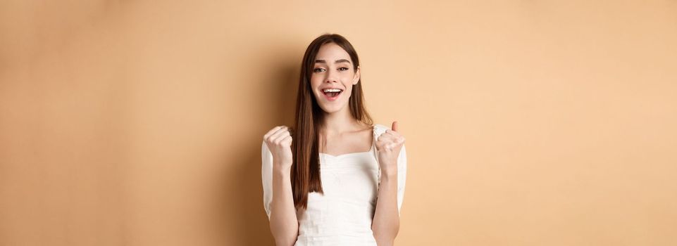 Happy smiling girl celebrating victory, making fist pump and say yes, achieve goal, triumphing or getting motivated, standing on beige background.