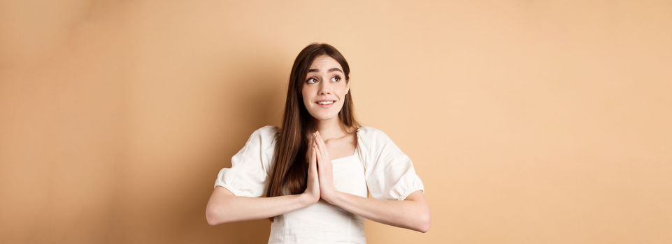 Hopeful young girl begging god and looking dreamy at upper left corner, praying or making wish, standing on beige background.