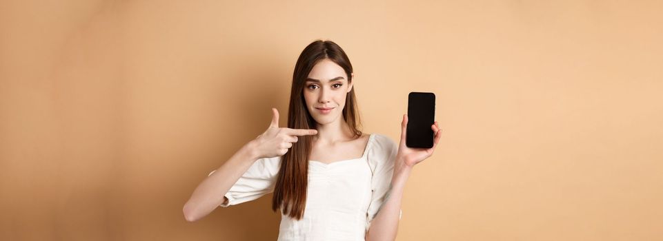 Confident girl pointing at empty smartphone screen, showing online promo deal, standing on beige background.